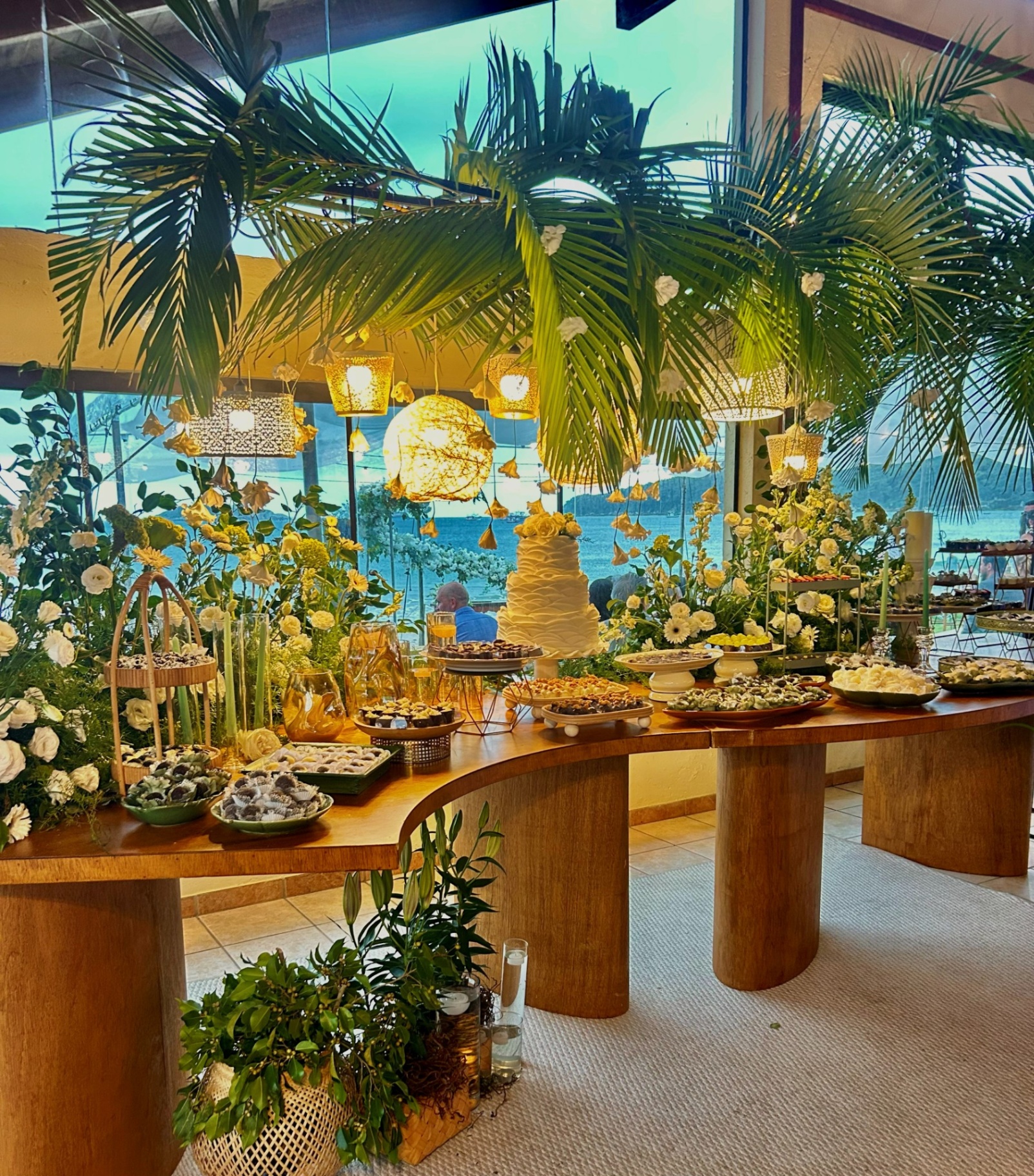 Mesa de doces tropical à beira-mar com flores brancas e amarelas e luminárias suspensas