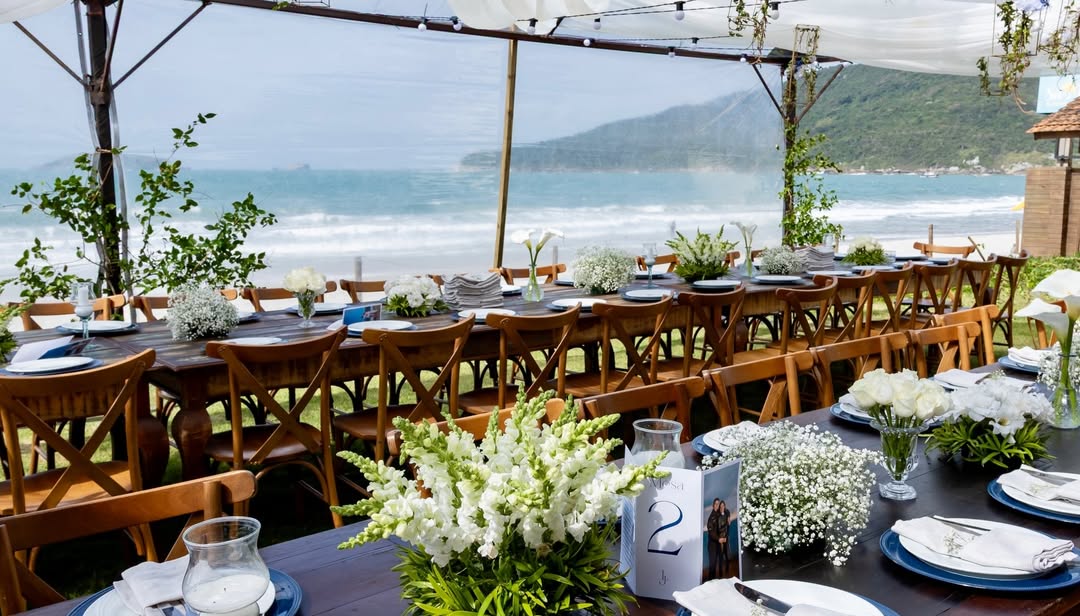 Mesas longas sob tenda branca com vista para o mar e flores brancas