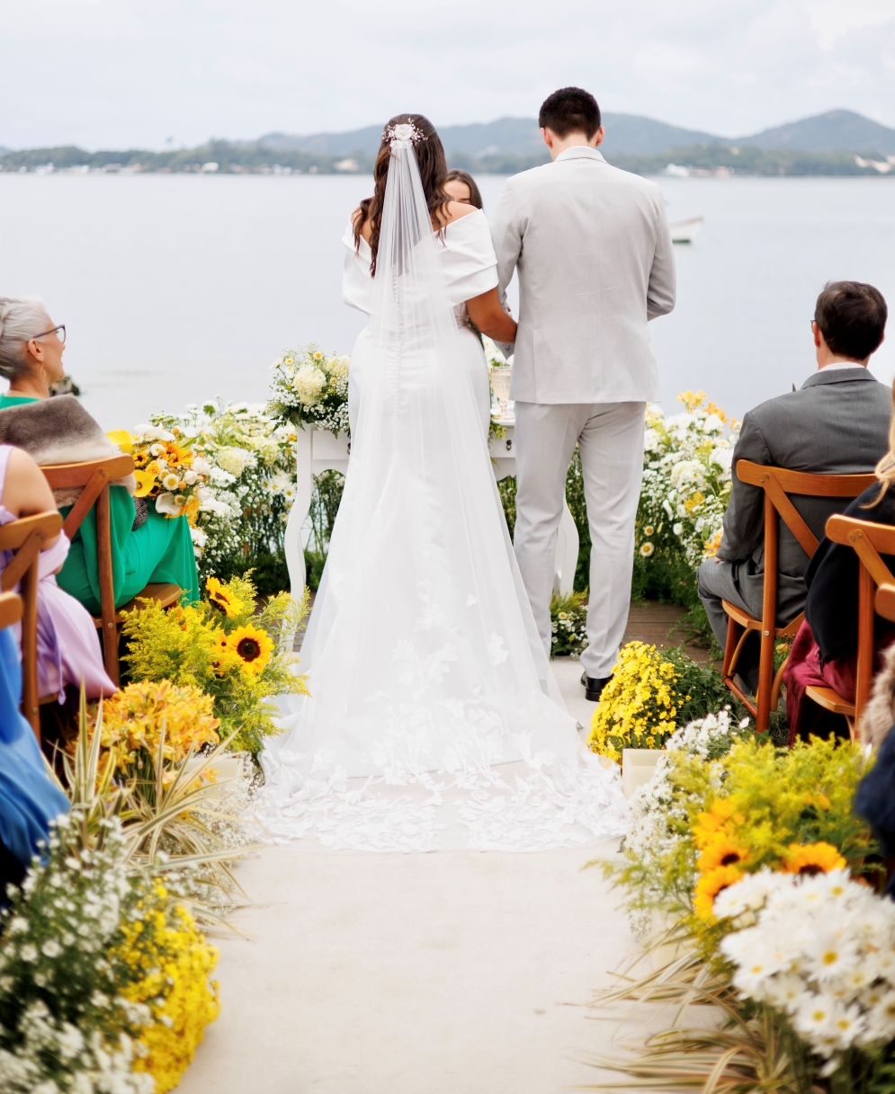 Cerimônia à beira da lagoa com noivos ao altar, aisle florido com girassóis e margaridas